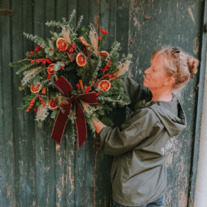 Woman holding a decorated Christmas wreath with berries, grasses, and orange slices in front of an old barn door