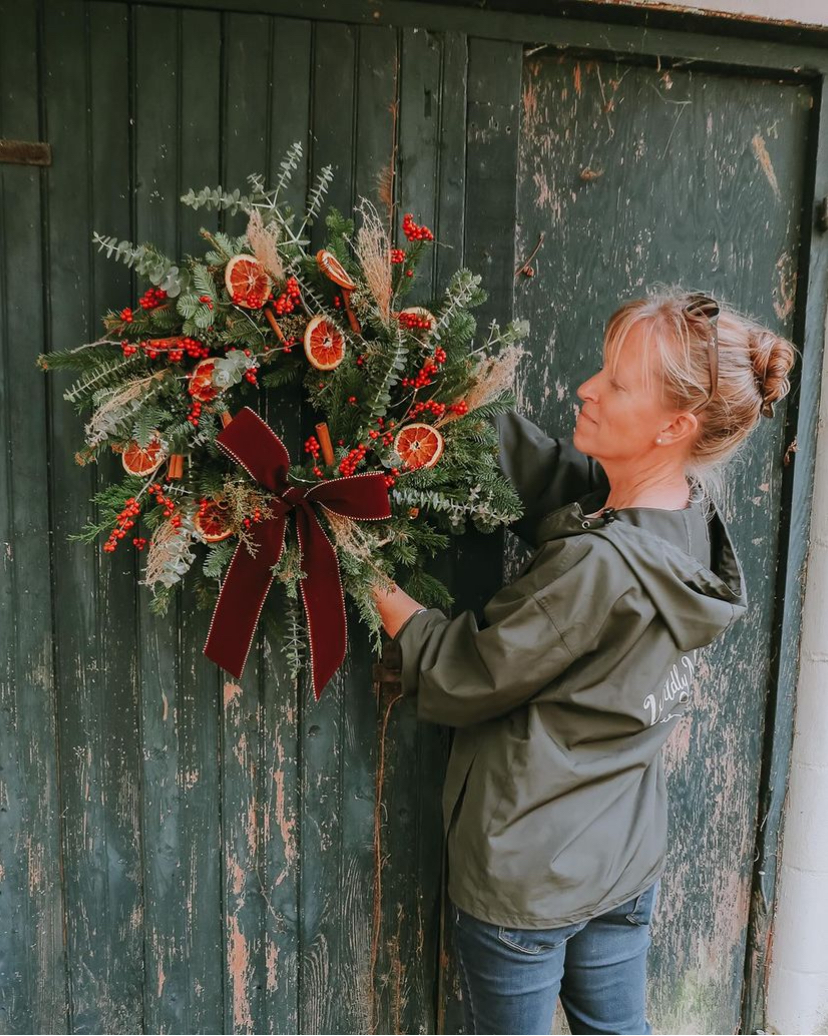 Woman holding a decorated Christmas wreath with berries, grasses, and orange slices in front of an old barn door