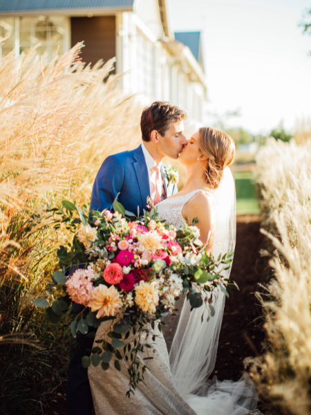 Bride kissing her groom amongst tall grasses holding an oversized cascading bouquet of yellow, and various pink flowers