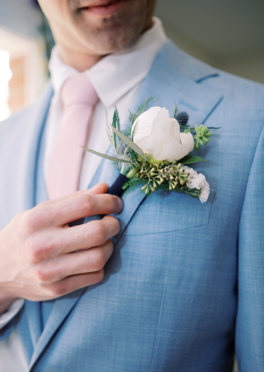 Blue suit with a pink tie, hand holding up a white floral boutonniere
