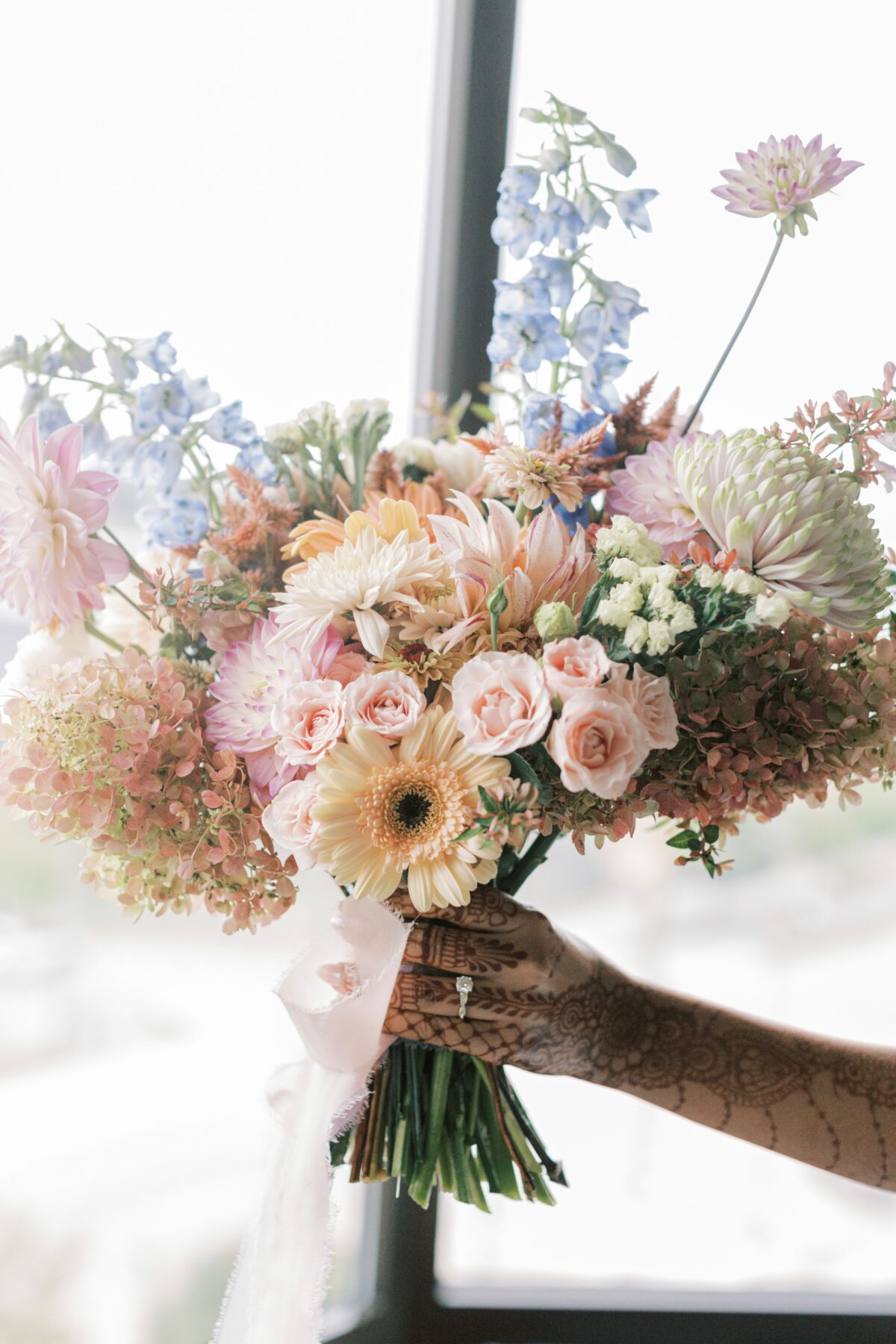 Bride holds bouquet of pastel summer florals in front of a window