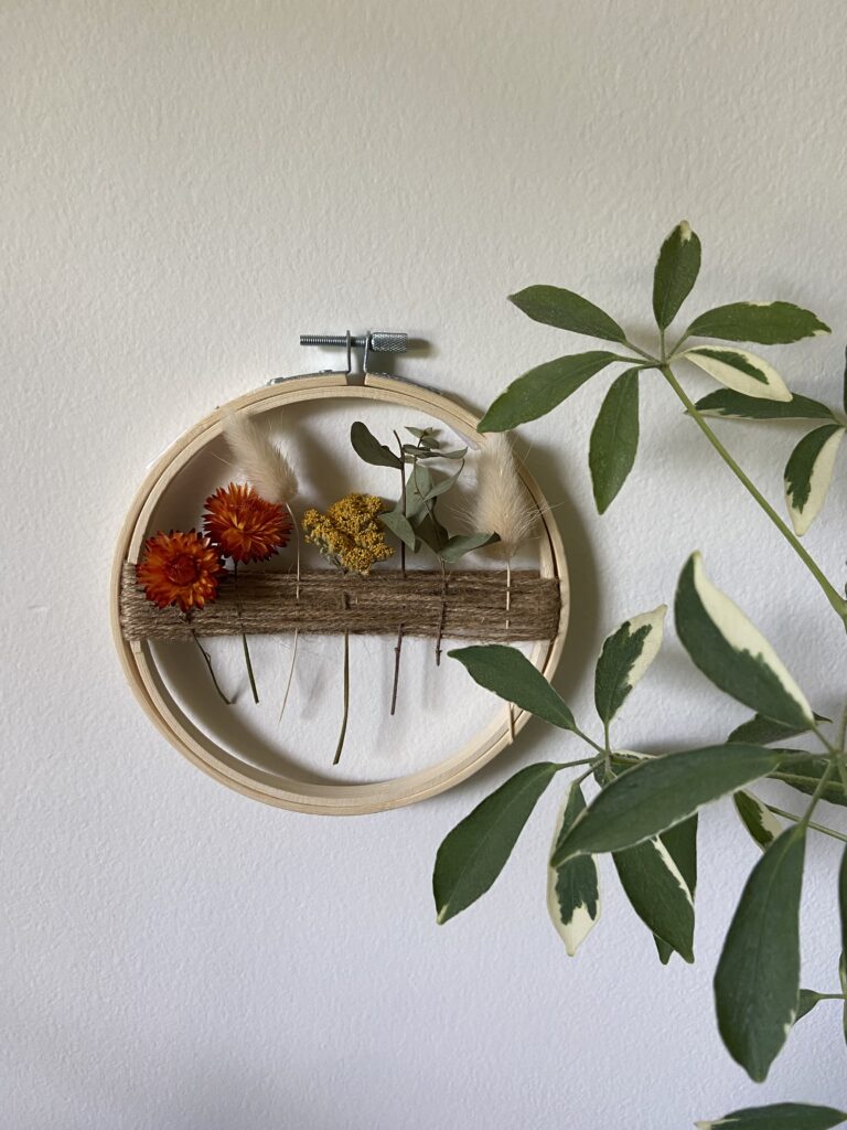 Embroidery hoop with twine and dried florals hung on a white wall with green indoor plant.