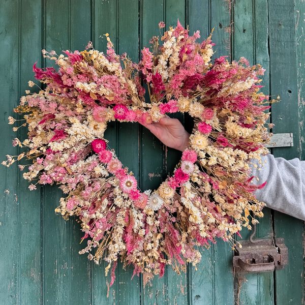 A pink and white dried flower wreath in a heart shape is held up in front of an old green barn door.