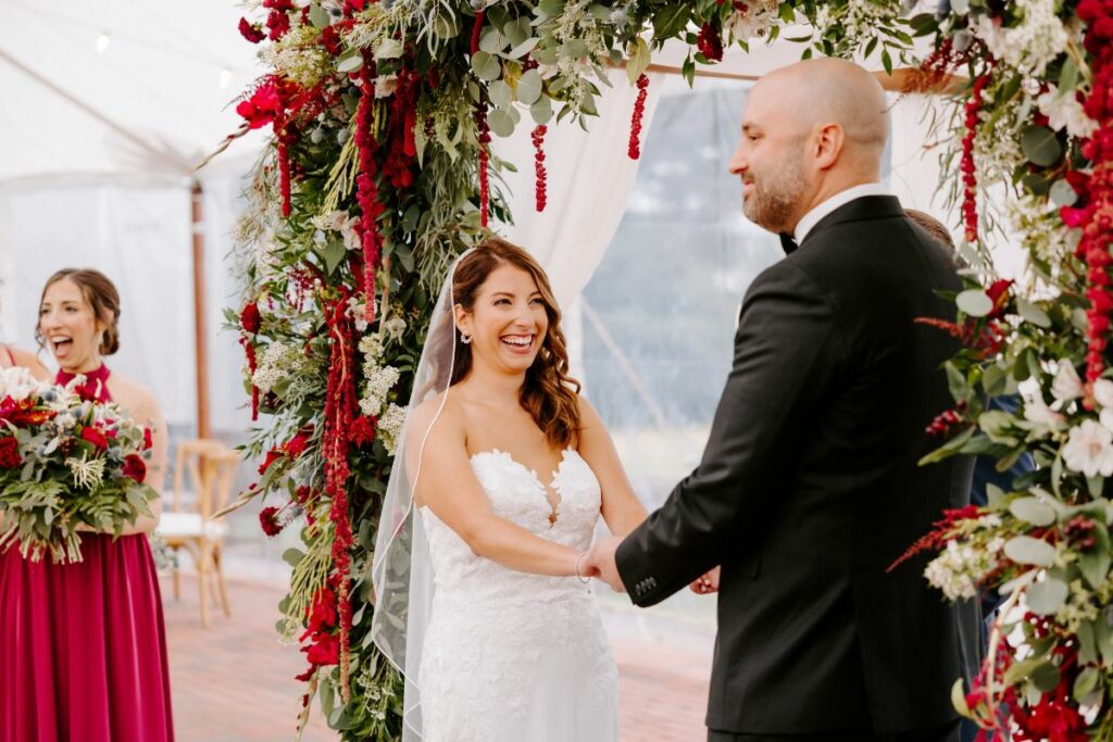 Bride and groom smiling during the ceremony, standing under the chuppah with thick jewel tone flowers.