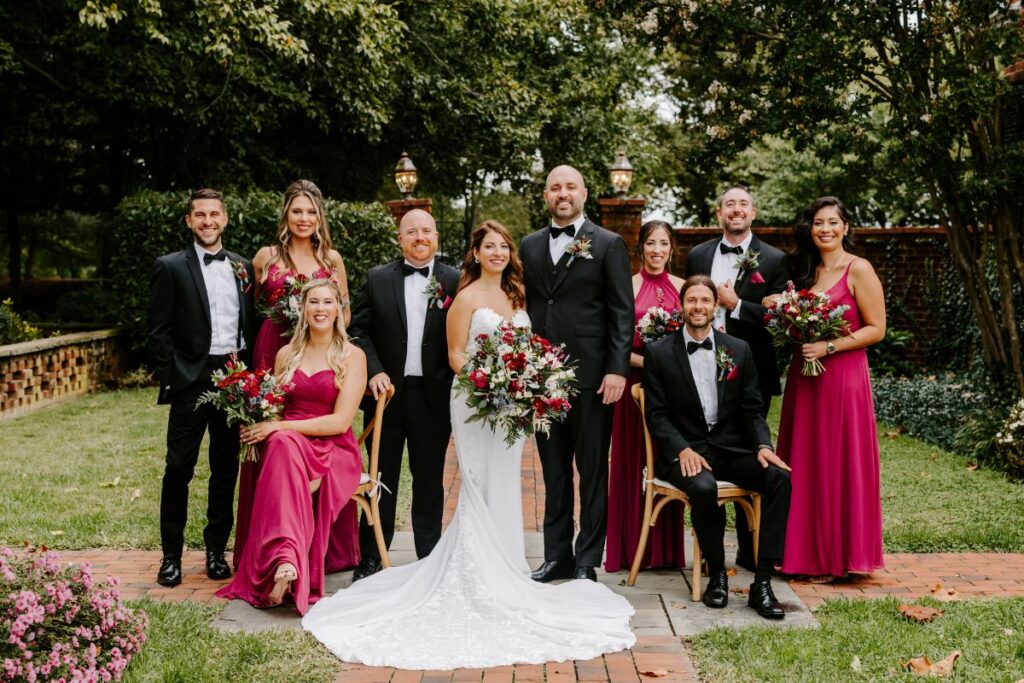 Full wedding party in black tuxedos and berry dresses gathered in the garden with jewel tone wedding flowers.