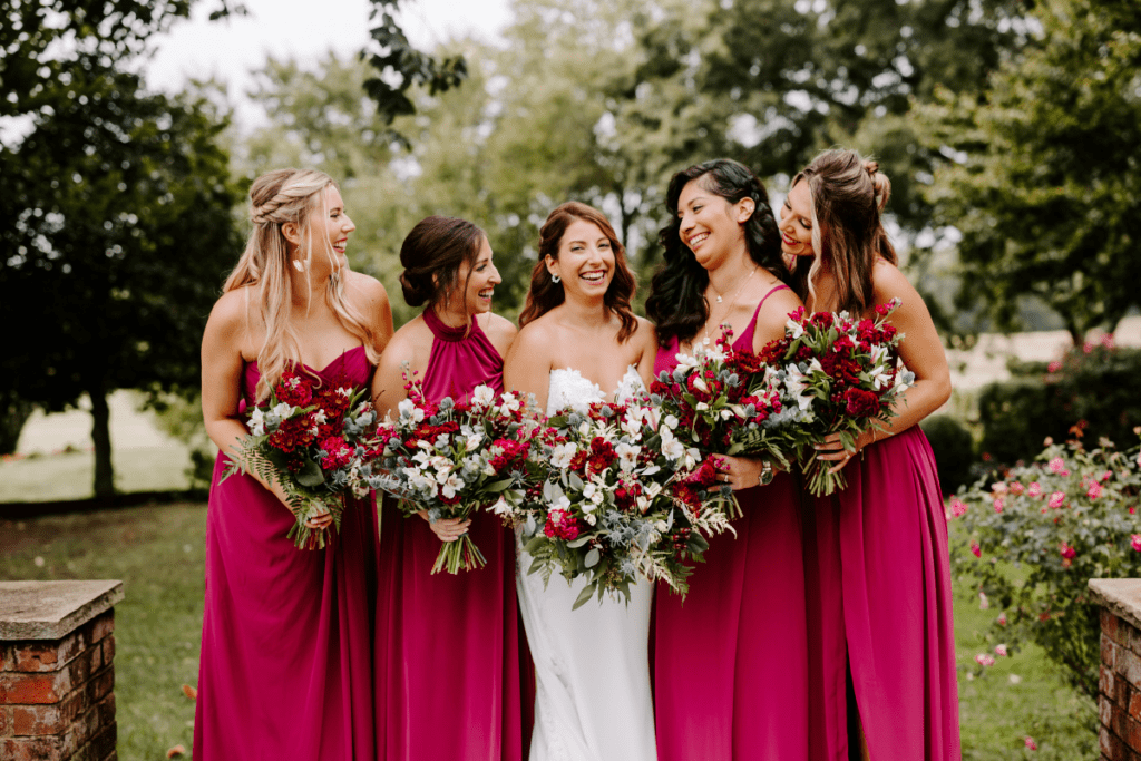 Bride and her bridesmaids smile in a group photo, wearing berry color dresses and holding flower bouquets in maroon jewel tones.