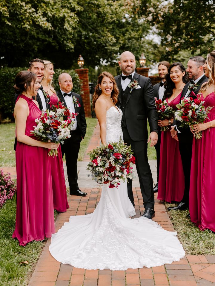 Bride and groom smile with the wedding party, wearing berry dresses and black suits with jewel tone wedding flowers.