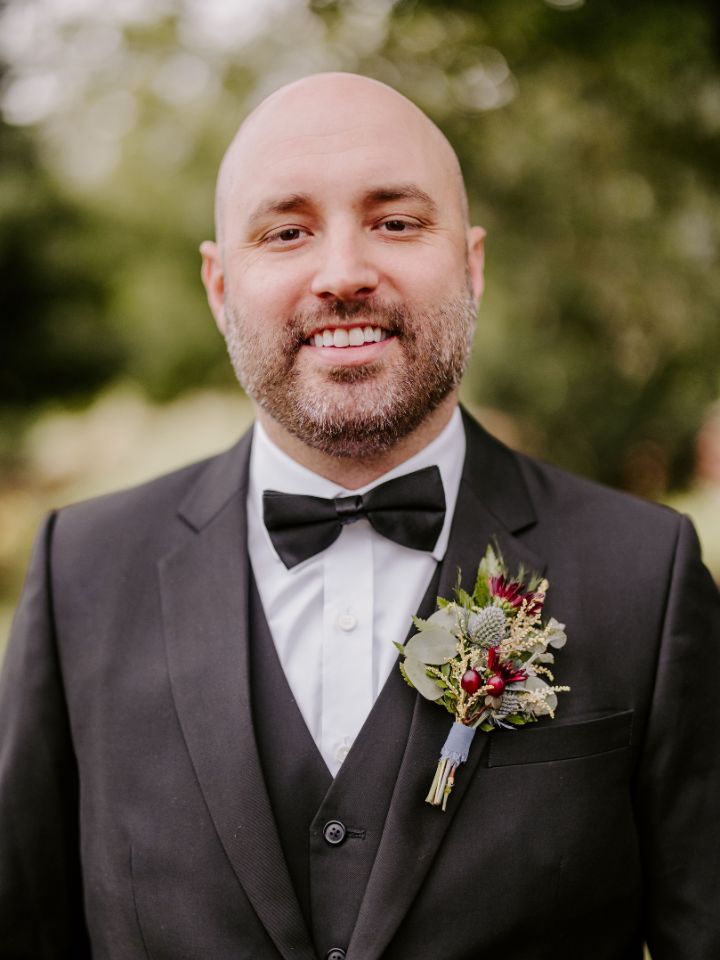 Groom smiles at camera in a black tux with bow tie, wearing a maroon and dusty blue flower boutonniere with eucalyptus.