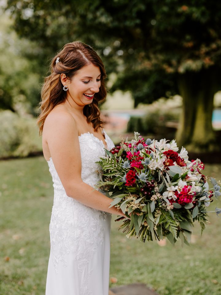 Bride smiles down at her bridal bouquet of fall jewel tone flowers in maroon and navy and white.