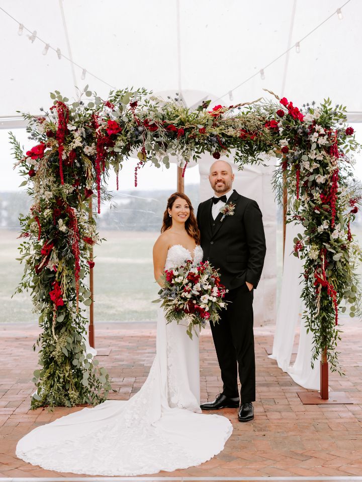 Bride and groom smile under the chuppah covered in maroon and dusty blue flowers.