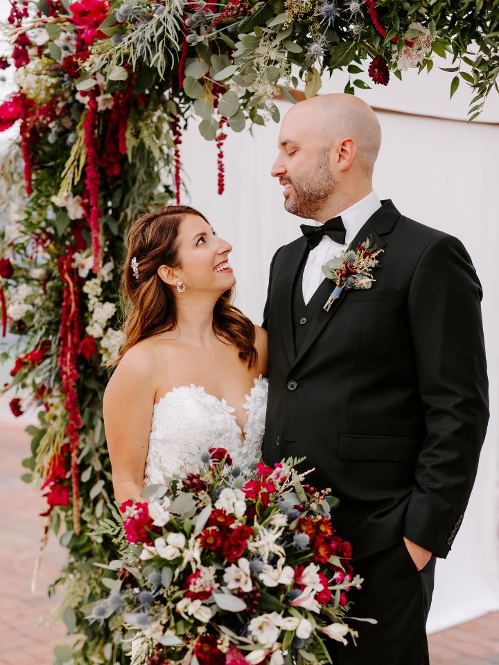 Bride and groom smile at each other underneath their jewel tone wedding flower chuppah.