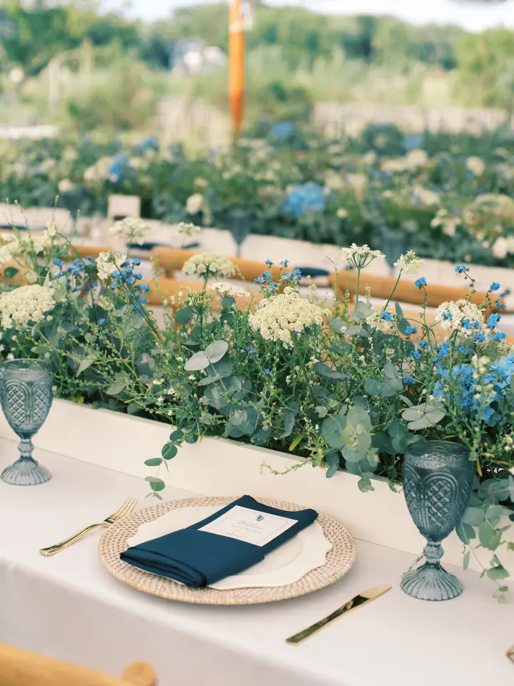 Long rectangle table with tan linen, beige charger and blue napkin with a greenery long arrangement