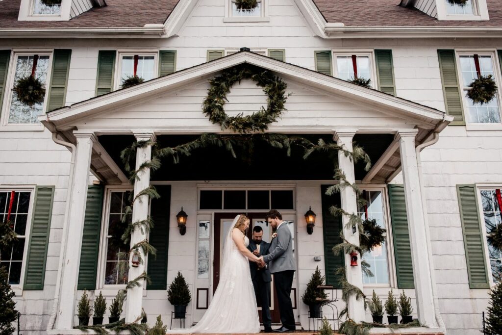Bride and Groom stand on front porch of an old Eastern Shore farmhouse for their wedding ceremony, with Christmas Wedding greenery, garlands, and wreaths.