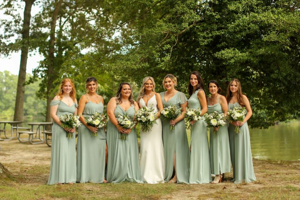 Bride and her bridesmaids wearing light sage green line up holding their white flower bouquets under the trees.