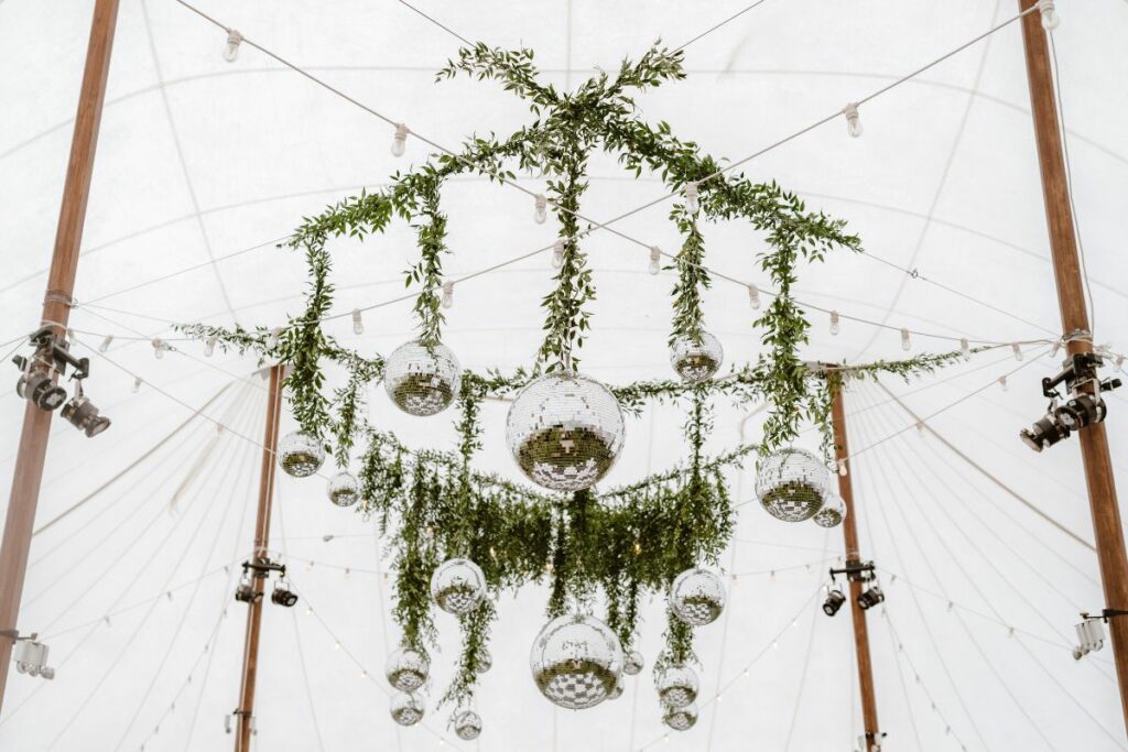 Looking up to the ceiling of a sailcloth tent, there is a large multi-disco ball installation with the wires covered in greenery.