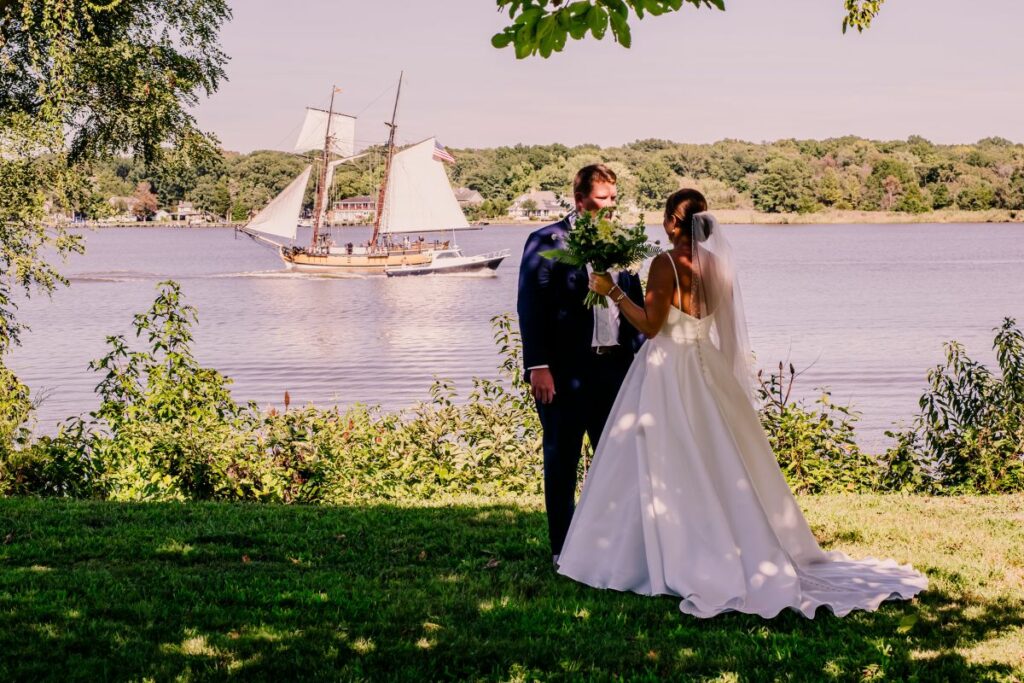 Bride and Groom stand onshore while a sailboat goes by in the background.