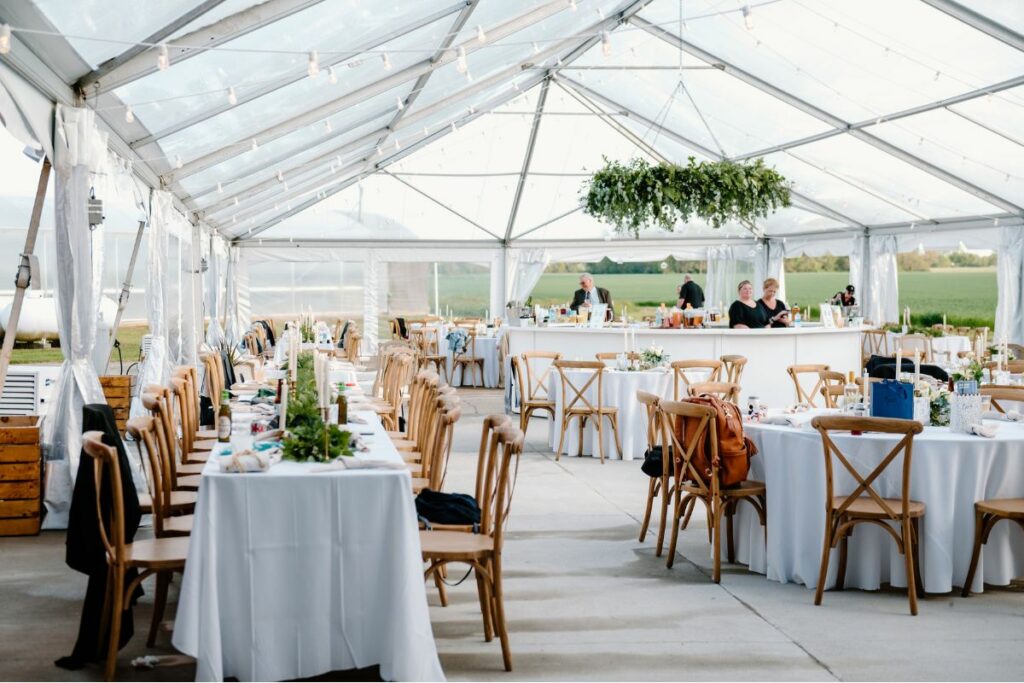 Clear top reception tent with guest tables in white linens, a round bar in the center, with a greenery chandelier.