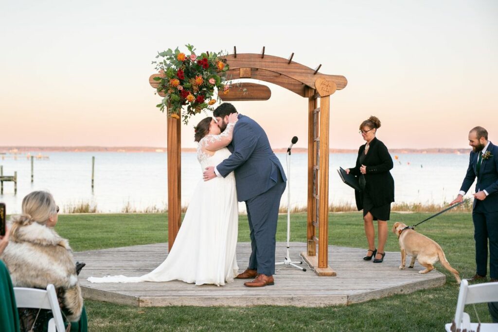 Bride and Groom kiss under ceremony arch during sunset in front of the water.