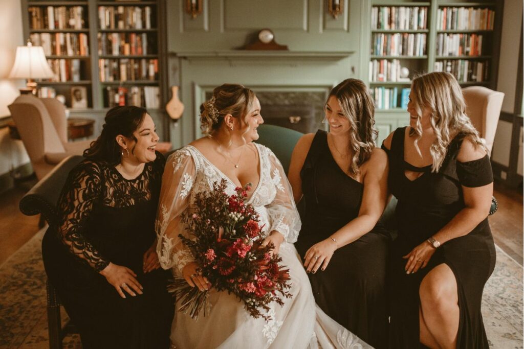 Bride and bridesmaids sit on a couch with black dresses and moody flowers.