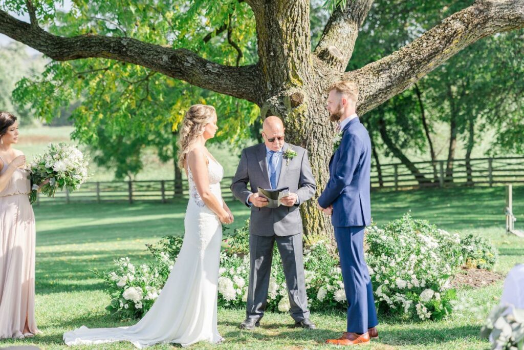 Bride and Groom stand under large tree with wedding officiant and circular ground arch of white and green wedding flowers.