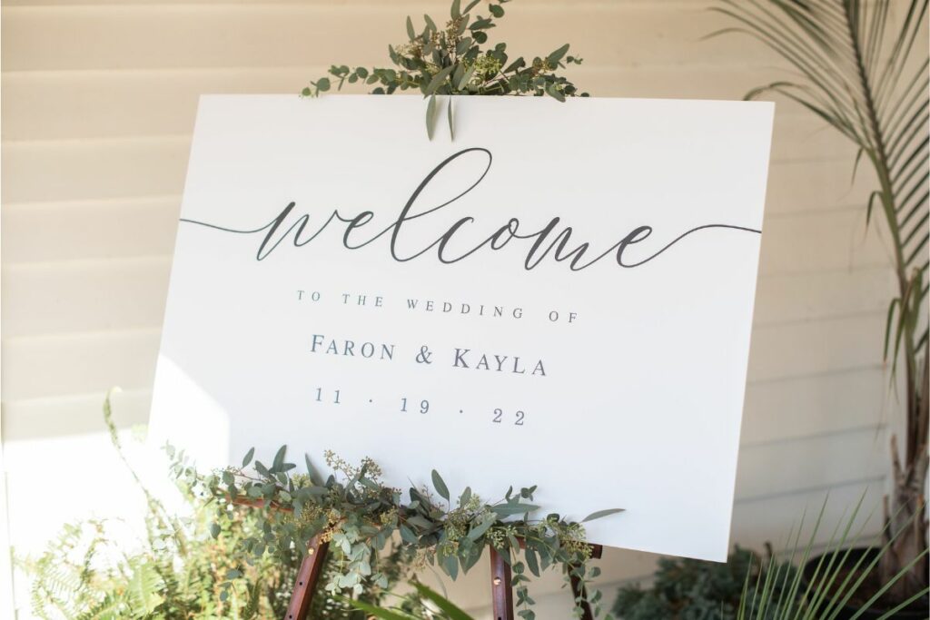 White welcome sign for a wedding with cursive script sits on an easel decorated with eucalyptus