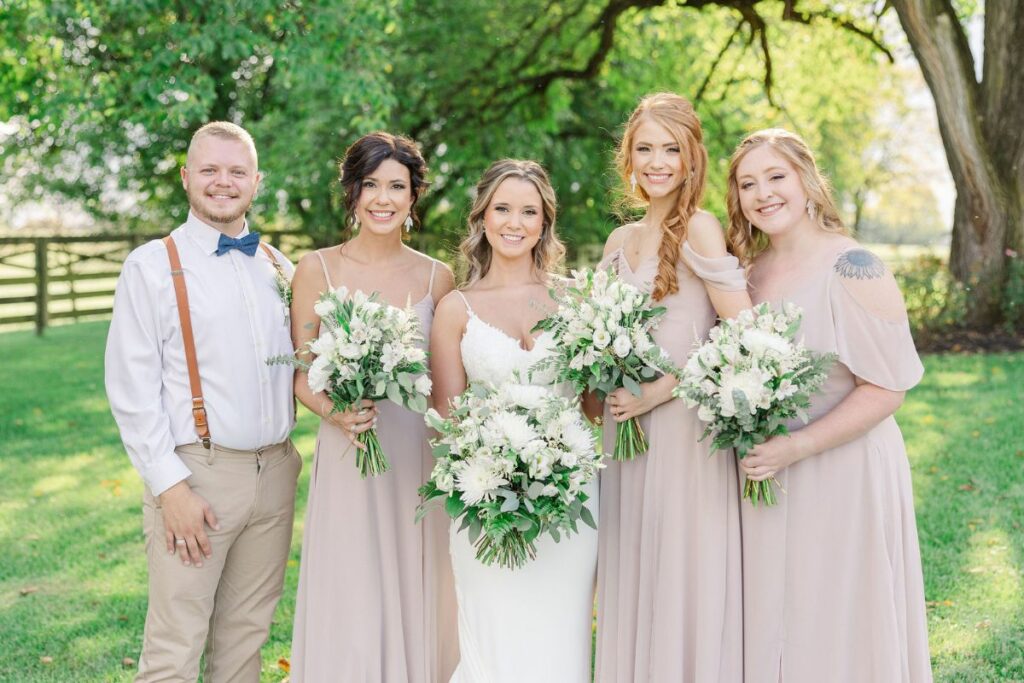Bride, bridesmaids, and bridesman smile at the camera outside with their wedding flowers.