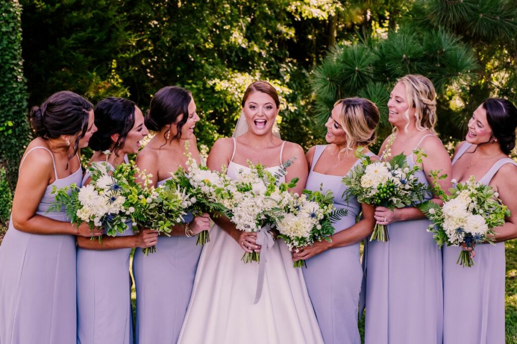 Bride has a huge smile in the middle of her bridesmaids, wearing blue and holding their floral bouquets.