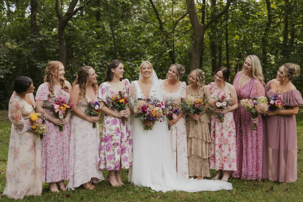 Bride and bridesmaids in floral print dresses lined up and smiling, holding their jewel tone wedding flowers.