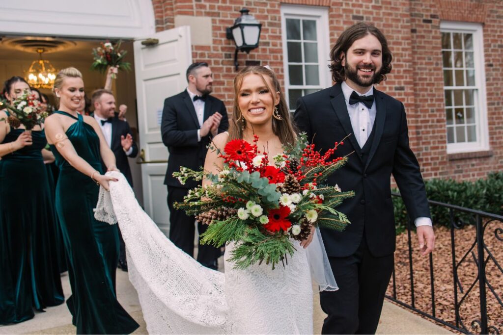 Bride and Groom leave church smiling after wedding ceremony, holding Christmas wedding flowers and surrounded by bridal party.