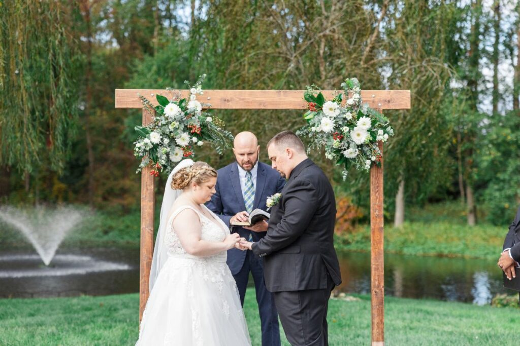Bride and Groom stand under a wooden arch with officiant for their wedding ceremony with flowers.