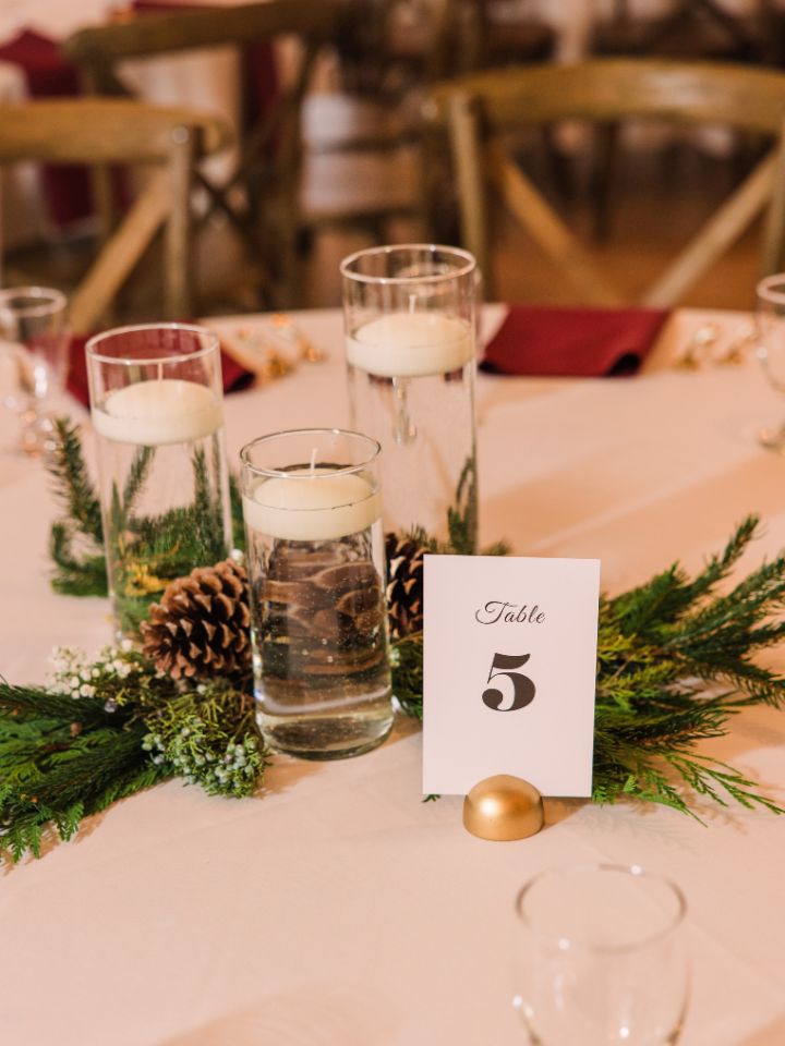 Guest table at a wedding reception has a white tablecloth, table number, glass cylinder trio with floating candles, and evergreen bundles.