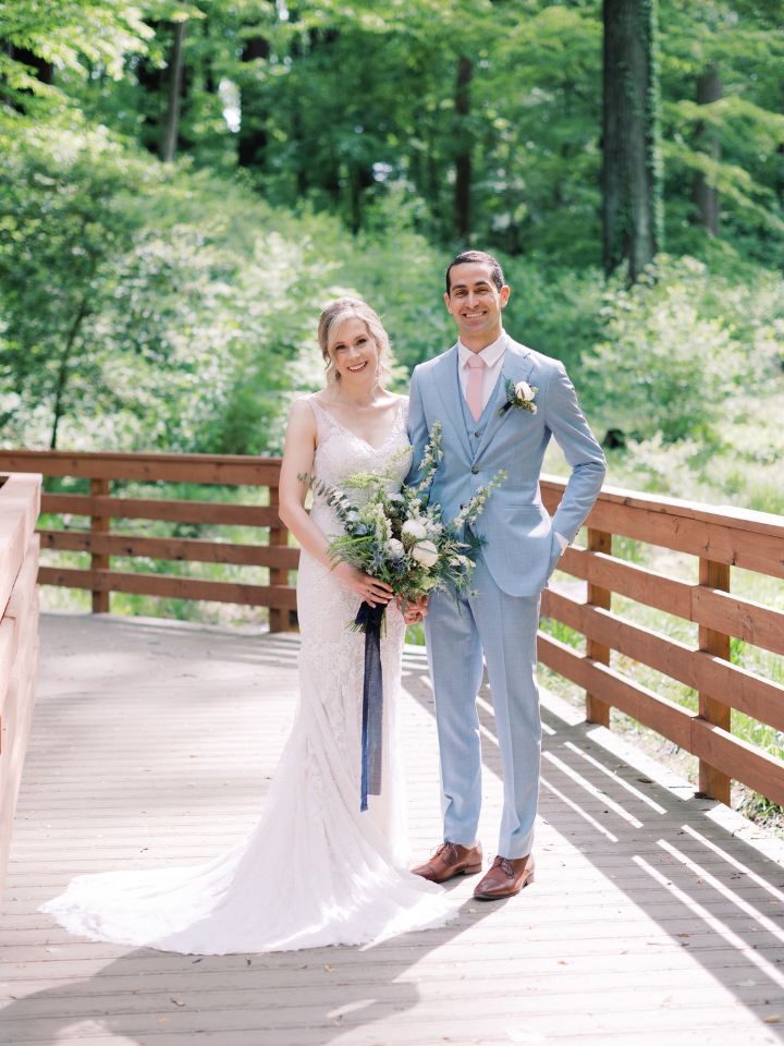 Bride and Groom pose on a wooden deck under the trees with white and blue wedding flowers.