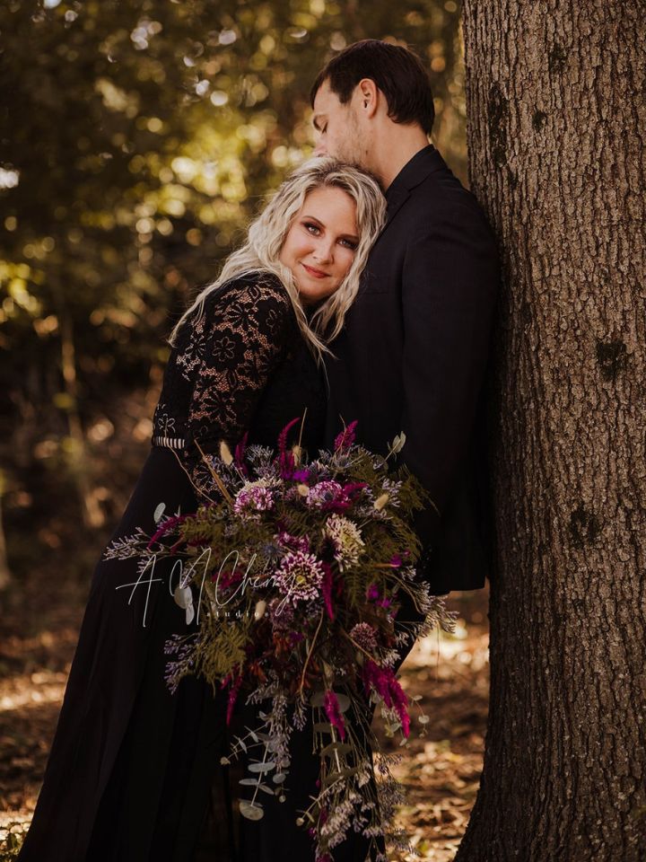 Couple leaning on tree holding a dark flower bouquet