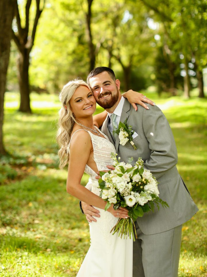 Bride and Groom smile at camera under the trees wearing a grey suit and white dress and holding white wedding flowers.