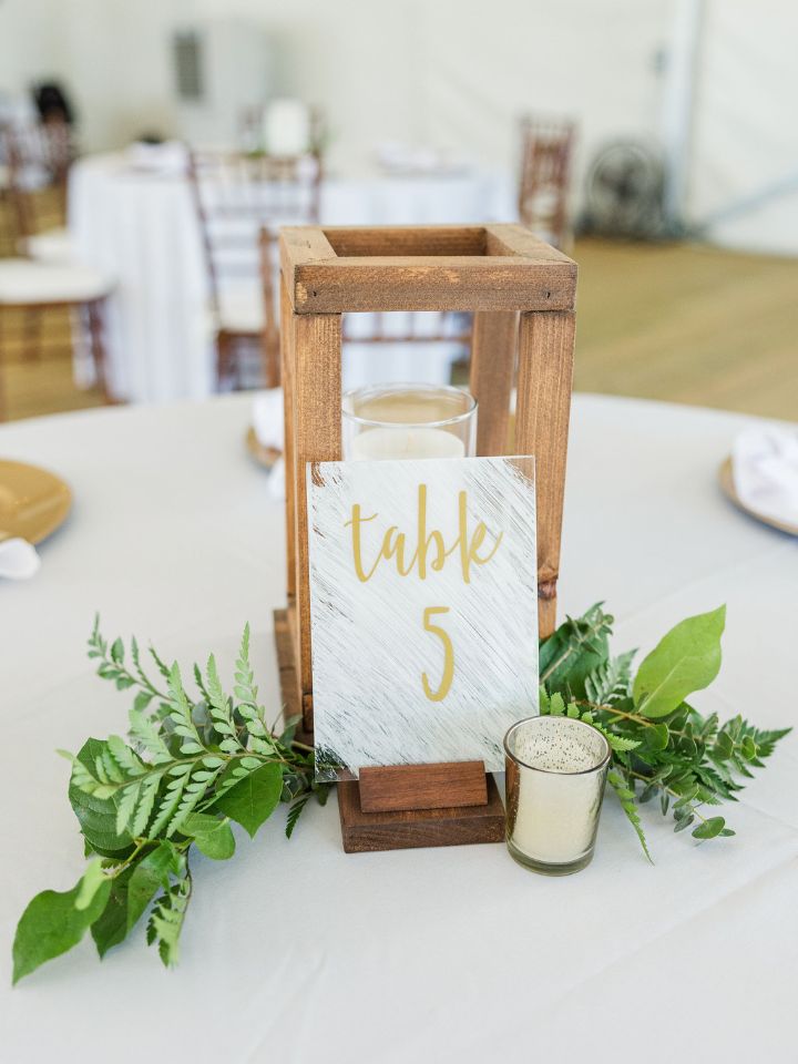 Wooden decorative lantern sits on a reception table with light greenery and a table number.