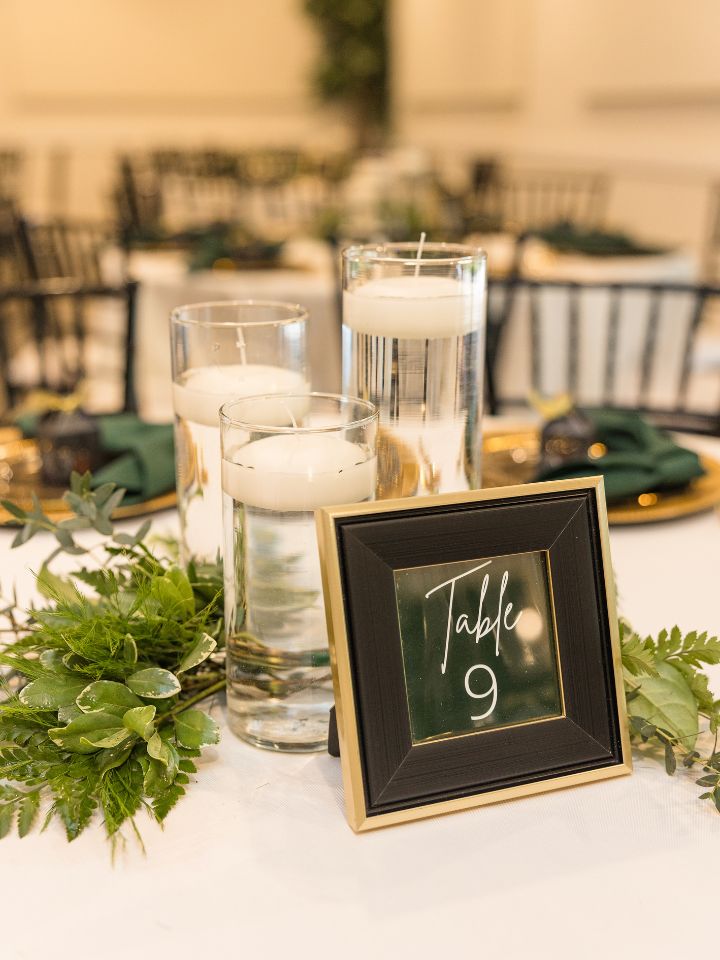Wedding reception table with white linen, trio of floating candles, and greenery with a black table number sign.