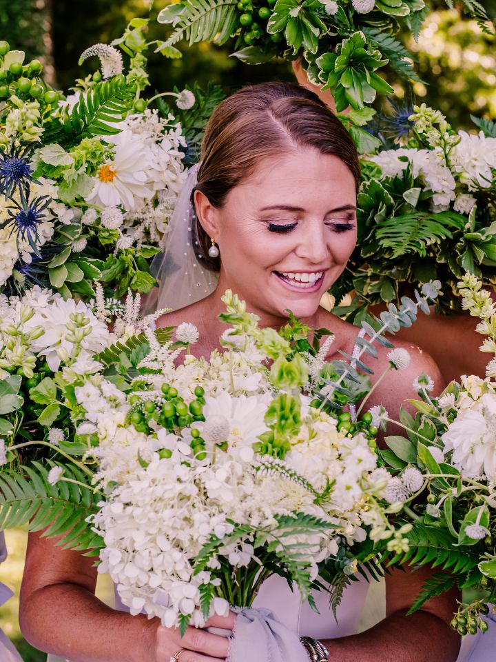 Bride smiles at her white and green wedding flowers, held around her face in a frame by her bridesmaids