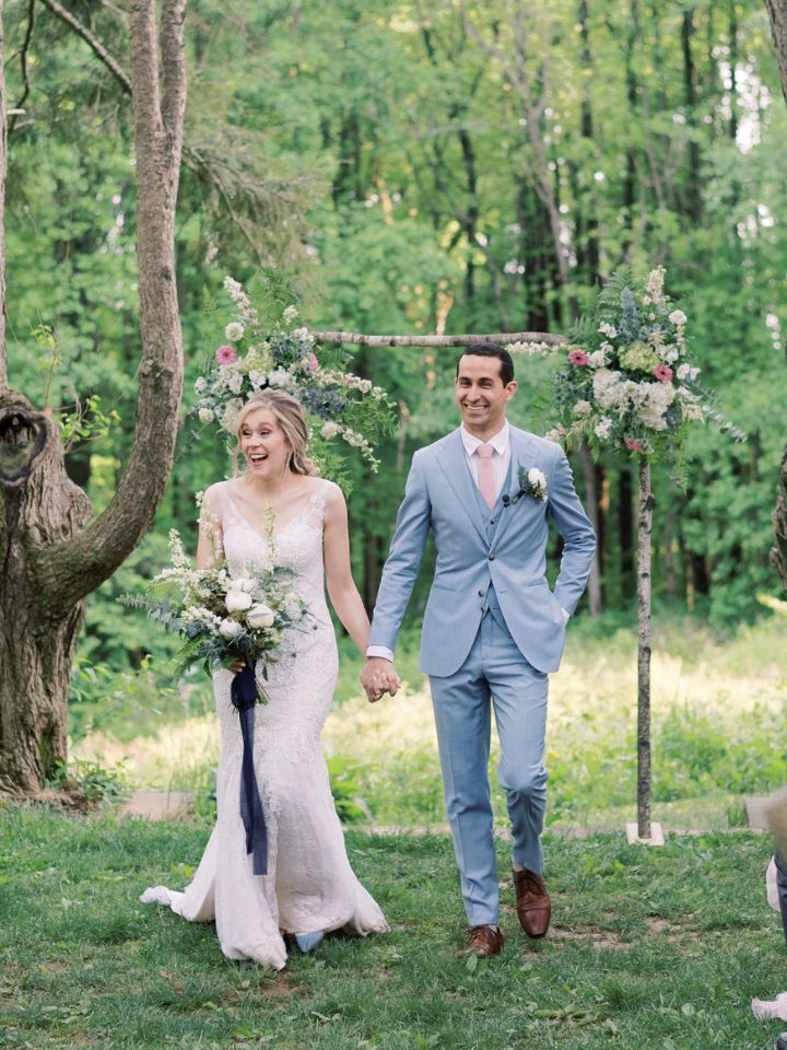 Bride and Groom walk back down the aisle holding wedding flowers under large trees.