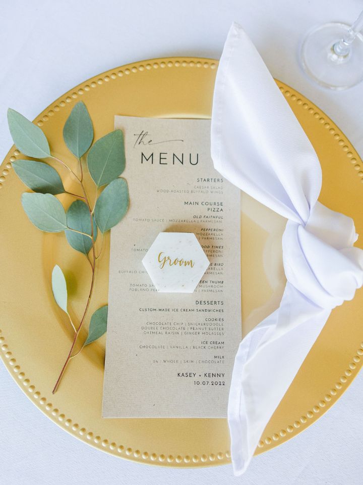 Gold charger on a white tablecloth has a knotted white napkin, menu, escort card, and single piece of eucalyptus.