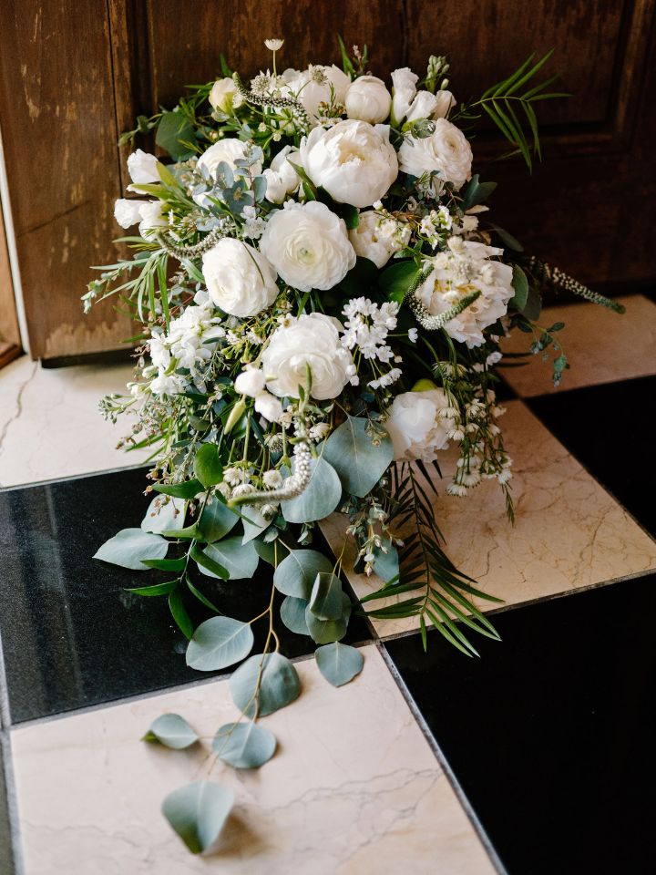 Cascading bridal bouquet in white and green wedding flowers sits on a black and white tile floor of a church.