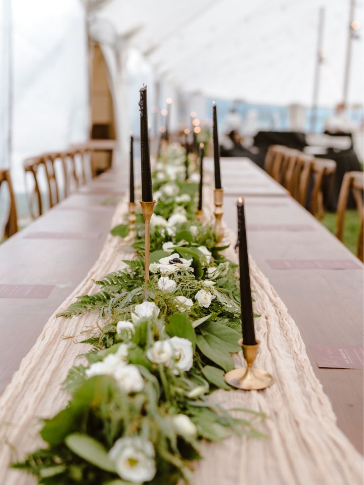 Wooden Harvest Table with muslin runner and greenery garland with white flowers, and black taper candles in gold holders.