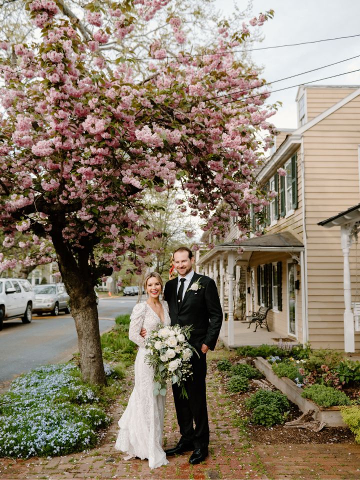 Bride and Groom stand under pink cherry tree holding white and green wedding flowers.