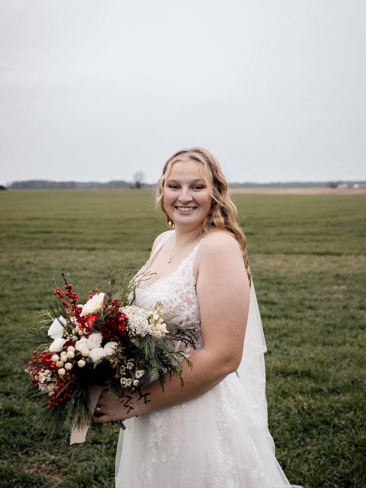 Bride smiles at the camera holding her Christmas wedding flower bouquet in red, white, and evergreens, outside near a field.
