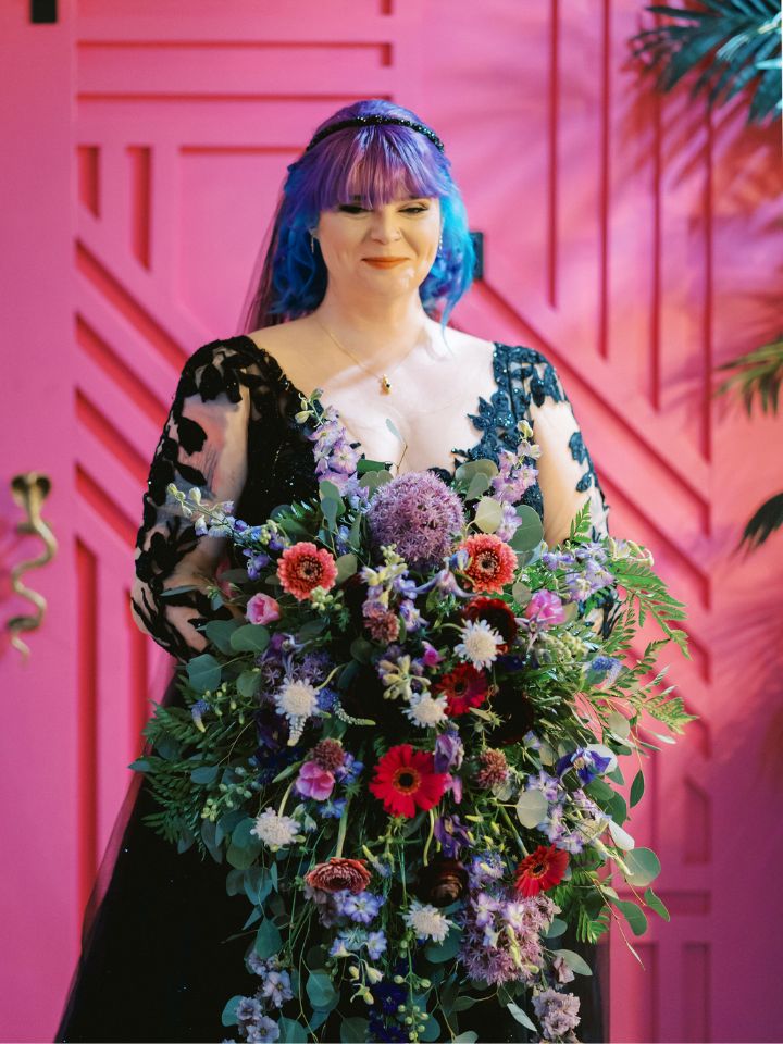 Bride wearing a black dress stands in front of a hot pink door holding a jewel tone flower bouquet.