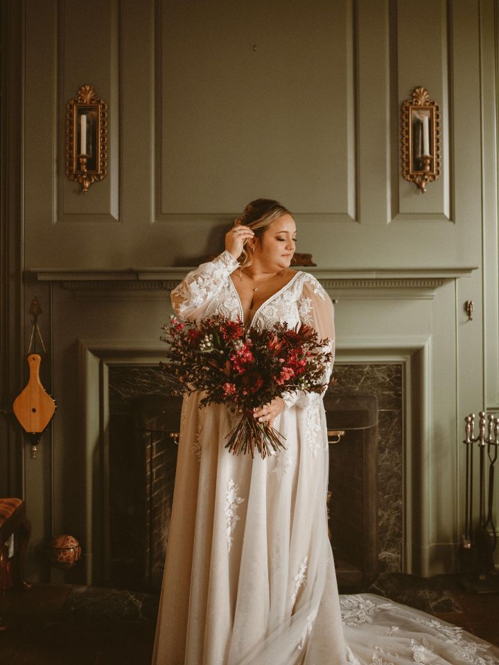 Bride holds wedding bouquet in moody jewel tone florals in front of an old fireplace.