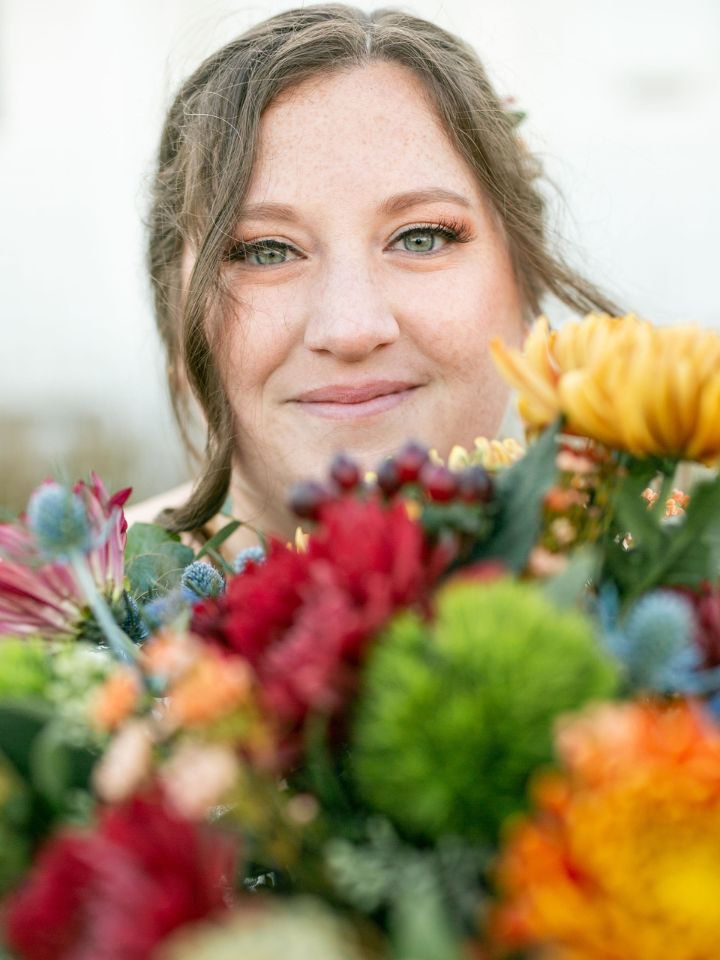 Bride looks at the camera from behind her bouquet of oranges, greens, burgundy, and navy blue.