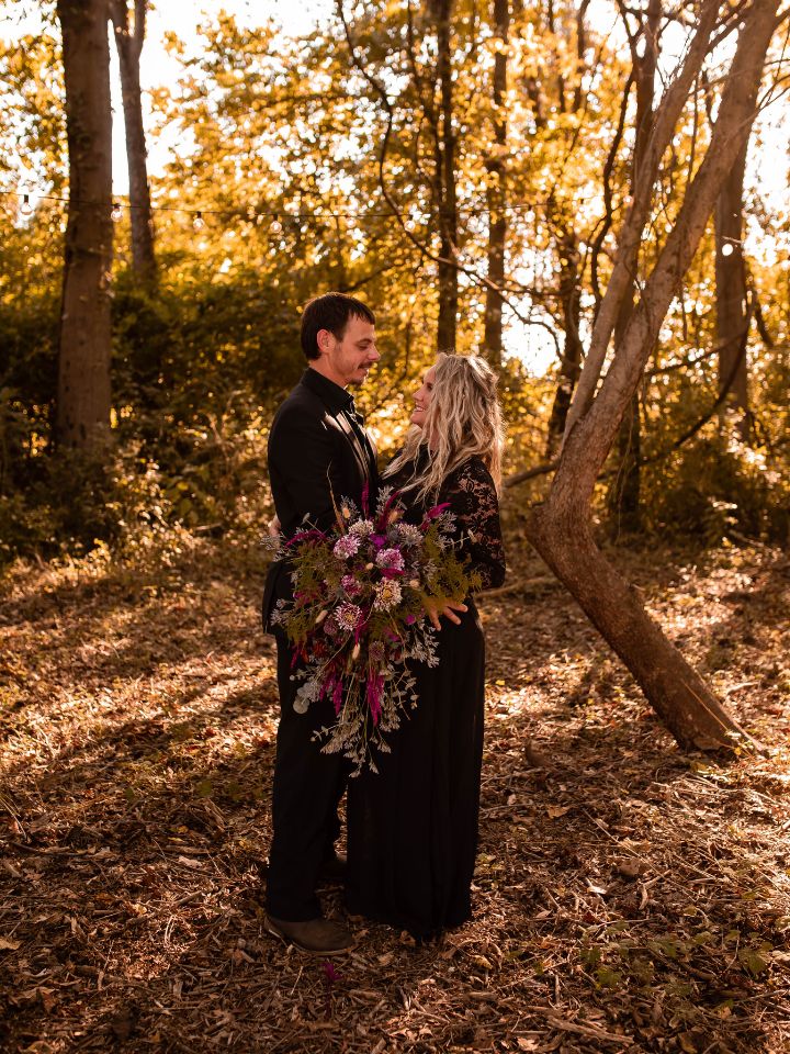 Couple standing in the woods holding a large flower bouquet.