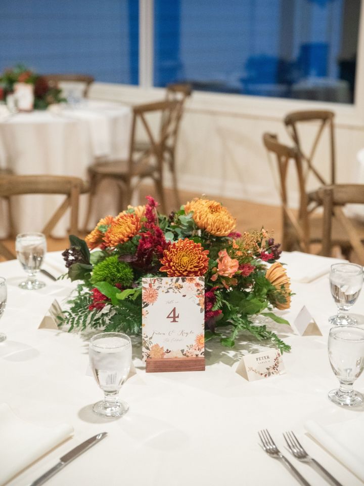 A guest table at a wedding reception has a white table cloth, table number, and low rounded arrangement of autumn wedding flowers