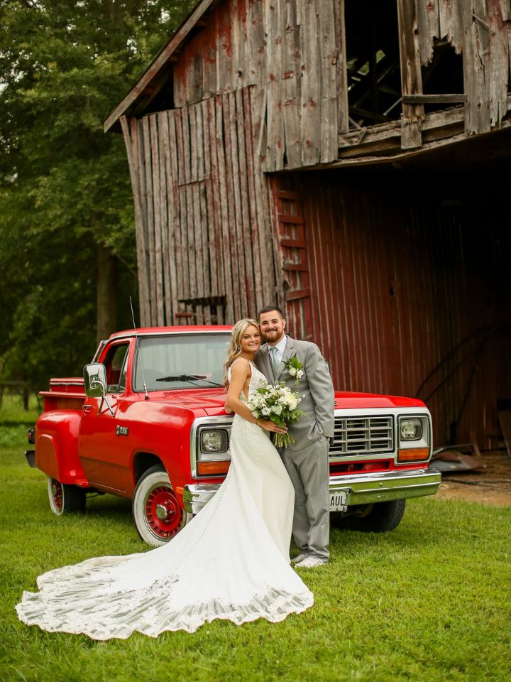 Bride and Groom stand in front of an old barn and a bright red truck, wearing a grey suit and white dress with white wedding flowers.