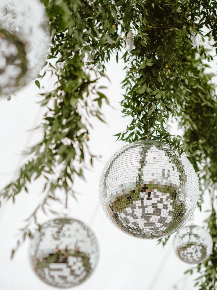 Close up of hanging disco ball installation from sail cloth tent ceiling, wires covered in greenery, and black and white dance floor reflected in ball.